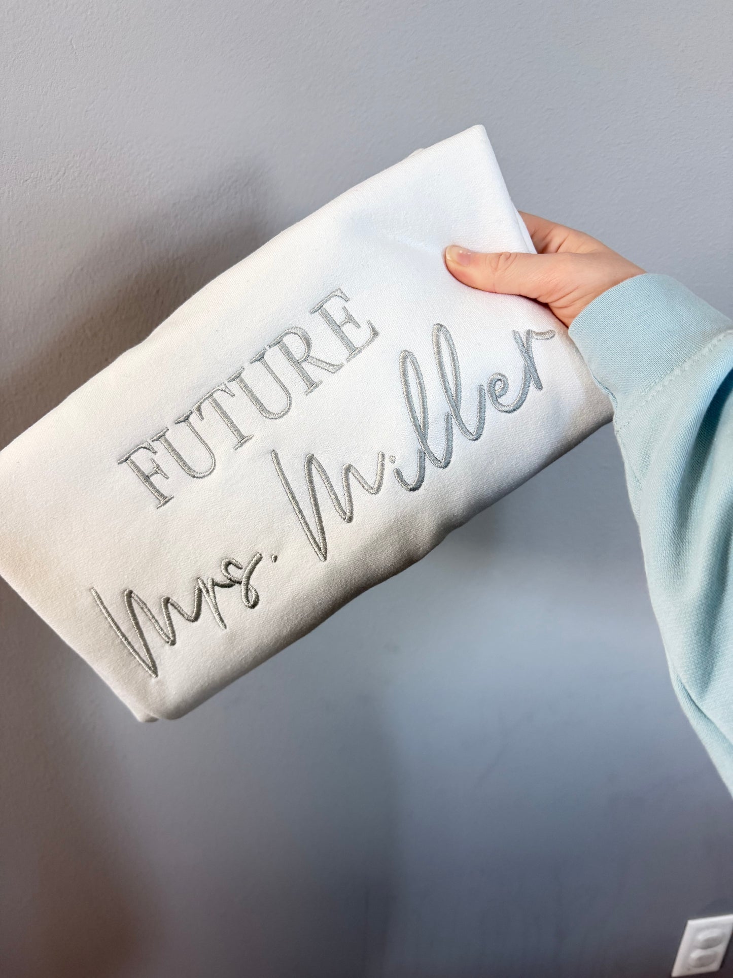 Person holding a white folded sweatshirt with 'FUTURE' and 'Mrs. Miller" text on a gray background