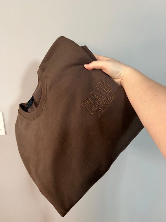 Brown sweatshirt folded and held up with "DAD" Athletic outline font and "EST. 2024" in brown thread.