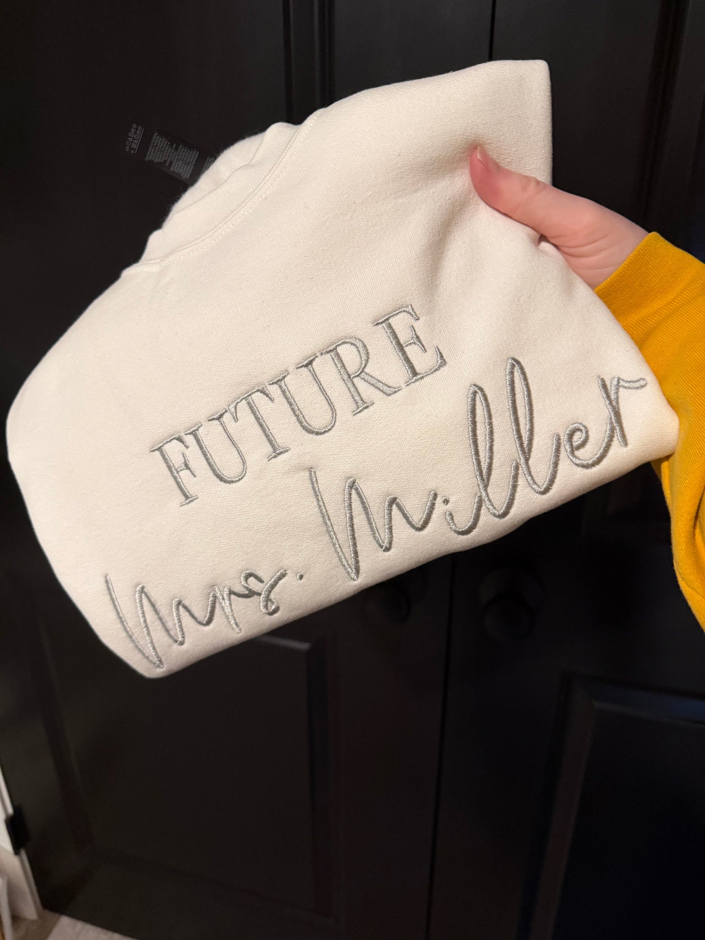 White folded sweatshirt with "FUTURE Mrs. Miller" in silver thread with dark background.