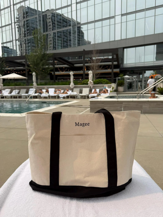 Beige tote bag with black handles and trim on a poolside table, with a pool and lounge chairs in the background.